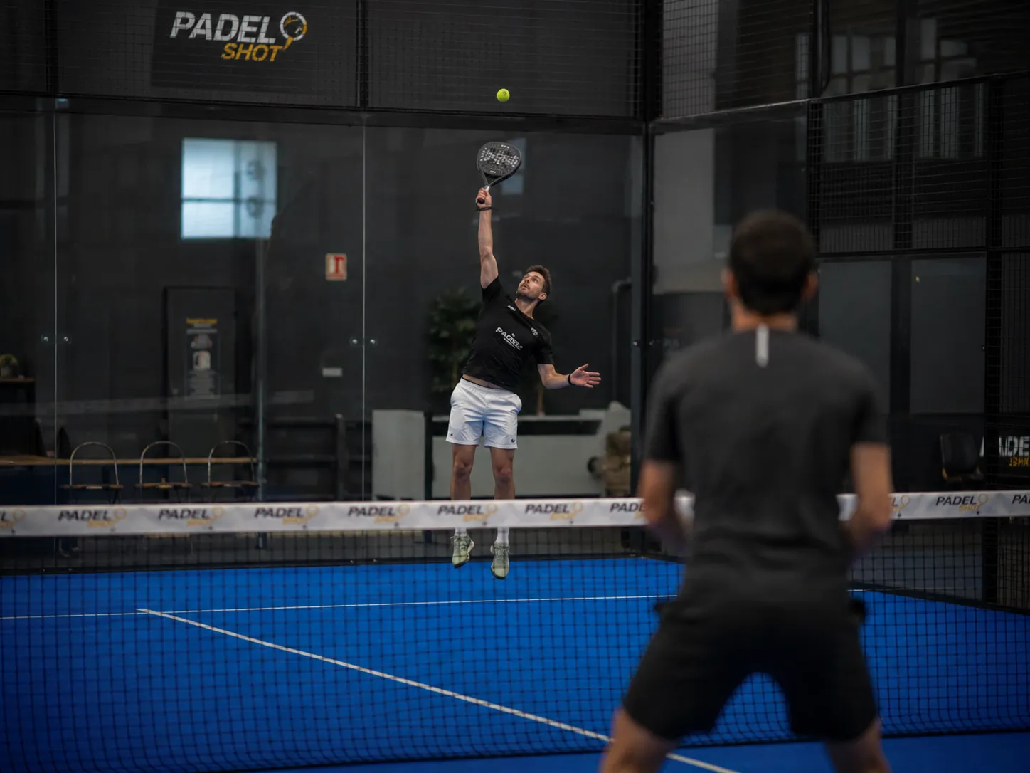 Player hitting a bandeja overhead, slightly back from the net, on a padel court